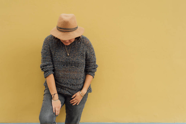 Donna irriconoscibile guarda in basso e si veste alla moda con un cappello e indossa vestiti lavorati a mano su una parete gialla. Moda di strada. Copia spazio - Foto, immagini