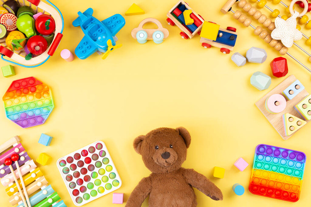 Baby-Kinderspielzeugrahmen mit Teddybär, Holz- und Musikspielzeug, Abakus, Flugzeug, Pop it Fidget Spielzeug und bunten Blöcken auf gelbem Hintergrund. Draufsicht, flache Lage - Foto, Bild