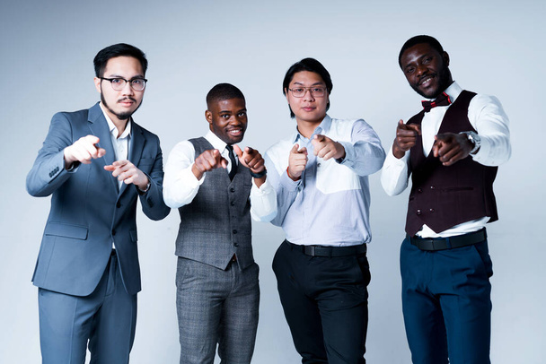African american and asian businessmen in suits smiling and pointing to the camera while standing on a light background - Foto, afbeelding