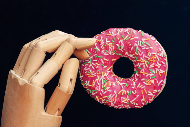 Close-up bright glazed donut and wooden robotic arm or manipulator on black background - Foto, Imagem