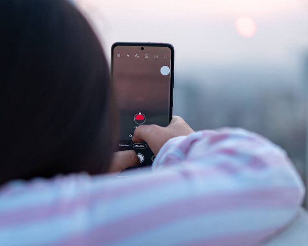 Молода дівчина брюнетка фотографує зі своїм телефоном на заході сонця
 - Фото, зображення