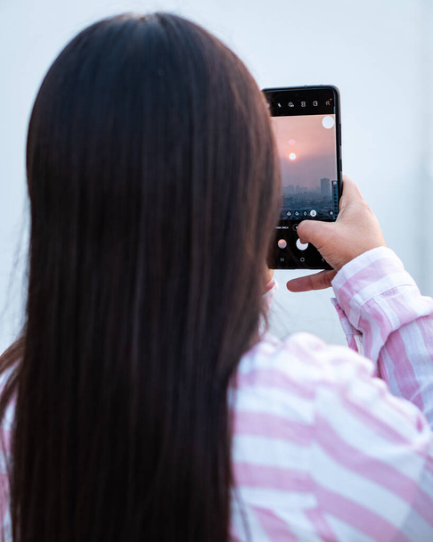 Молодая брюнетка, фотографирующая на закате на телефон - Фото, изображение