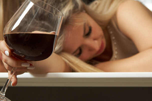 Wine glass on the background of a pretty tipsy blonde woman in an evening dress. - Zdjęcie, obraz