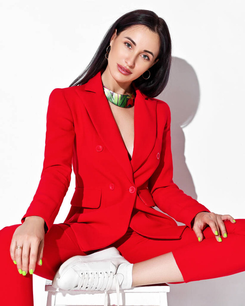 Daring brunette woman in red official pantsuit, jacket with deep neckline on naked body and sneakers sits on a stool - Photo, Image