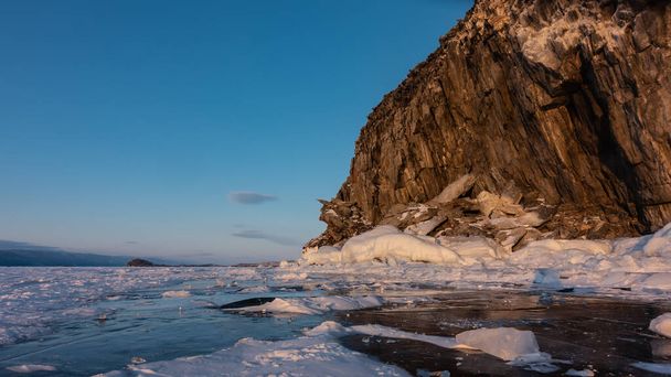 A whimsical rock, devoid of vegetation, against the backdrop of a blue sky. Snow on sheer granite slopes and on the ice of a frozen lake. Reflection on the surface. Evening golden hour. Baikal - Photo, Image