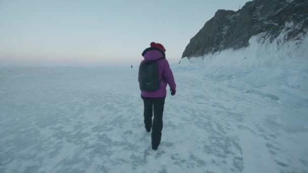 Una joven camina a lo largo del congelado lago Baikal pasando por bloques de hielo y montañas rocosas en la orilla. Vista trasera. Movimiento lento - Imágenes, Vídeo