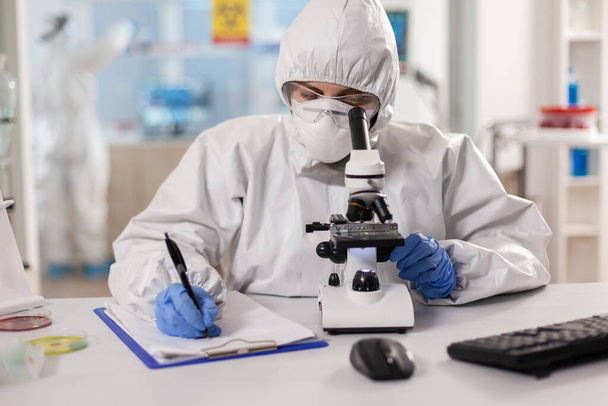 Científico de la medicina vestido con traje de ppe tomando notas durante la investigación de vacunas - Foto, Imagen