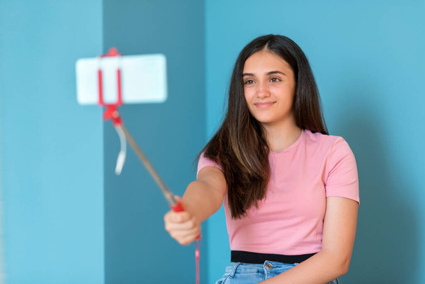 Menina adolescente bonita sorrindo ao tirar fotos selfie com seu telefone celular para mídias sociais e comunicação online contra a parede azul para espaço de cópia - Foto, Imagem
