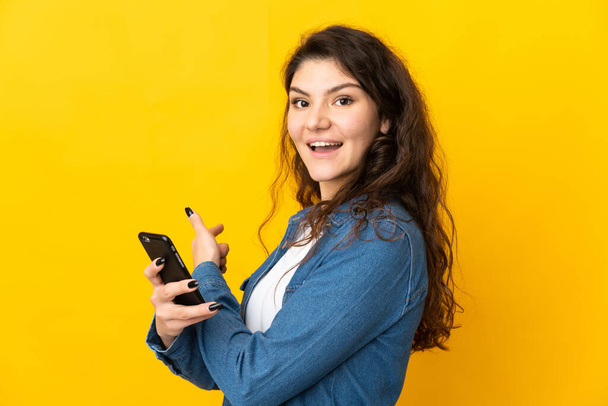 十代のロシア人の女の子は、携帯電話を使用して黄色の背景に隔離され、後ろを指す - 写真・画像