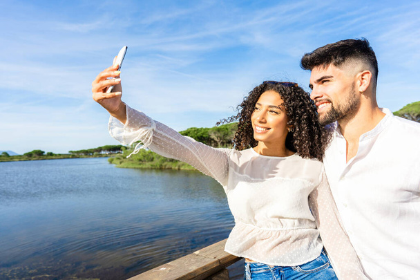 Счастливая пара в любви делает селфи на отдыхе на морском курорте на закате. Молодая афро-американка использует смартфон для автопортрета со своим парнем на озере. Миллениалы - Фото, изображение