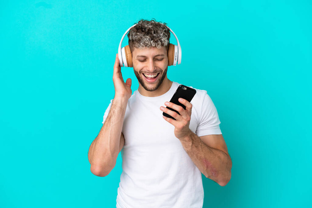 Fiatal jóképű kaukázusi férfi elszigetelt kék háttér hallgat zenét egy mobil és énekel - Fotó, kép