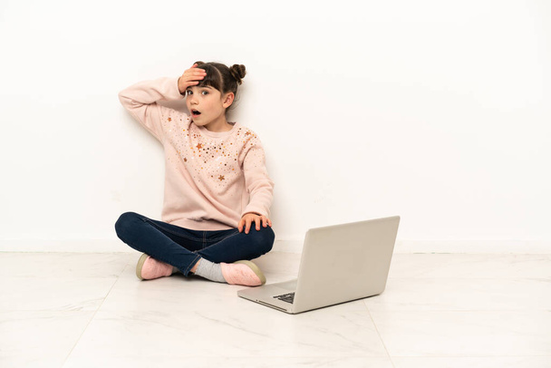 Dizüstü bilgisayarı olan küçük bir kız yerde oturuyor ve yan tarafa bakarken sürpriz jest yapıyor. - Fotoğraf, Görsel