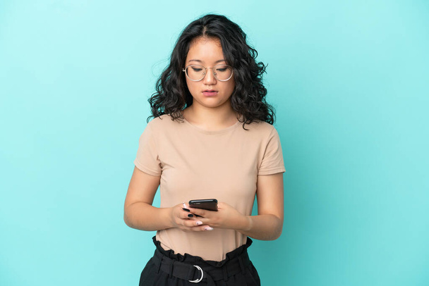 Mavi arka planda izole edilmiş genç Asyalı kadın cep telefonuyla mesaj gönderiyor. - Fotoğraf, Görsel