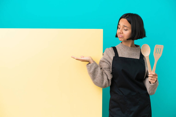 Jeune cuisinière métisse avec une grande pancarte isolée sur fond bleu tenant imaginaire copyspace sur la paume pour insérer une annonce - Photo, image