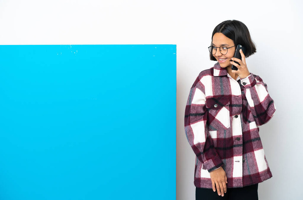 Young mixed race woman with a big blue placard isolated on white background keeping a conversation with the mobile phone with someone - Photo, Image