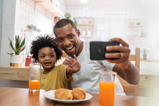 Happy Smiling African American Father and little son doing video call with smartphone and waving hands to their family in kitchen. Black family having fun while eating breakfast at home. - Fotoğraf, Görsel