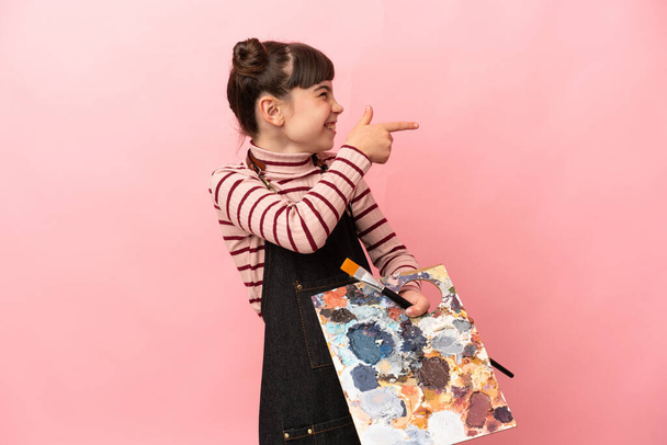 Malá umělkyně drží paletu izolovanou na růžovém pozadí ukazující prstem na stranu a prezentující produkt - Fotografie, Obrázek