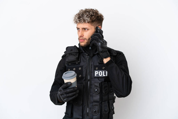 SWAT caucasian man isolated on white background holding coffee to take away and a mobile - Фото, изображение