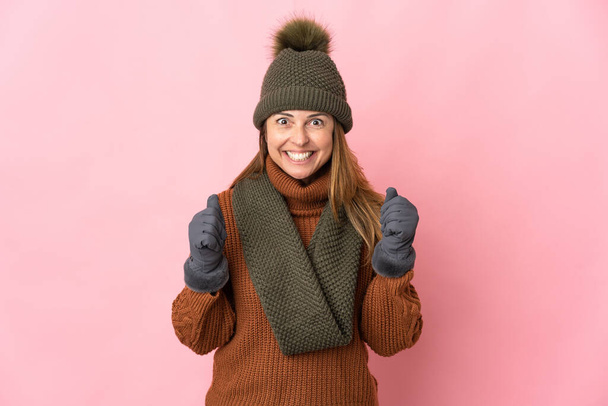 Middle age woman with winter hat isolated on pink background celebrating a victory in winner position - Photo, Image