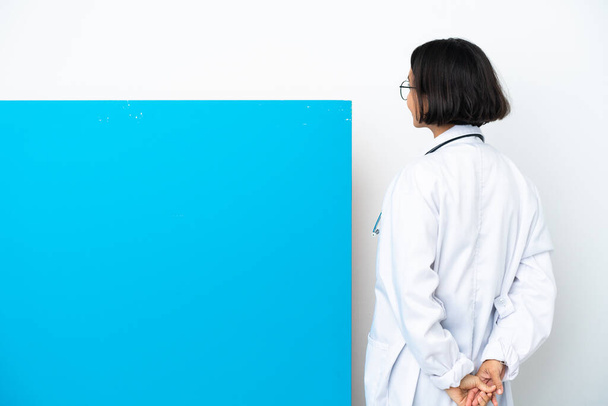 Joven doctora de raza mixta con un gran cartel aislado sobre fondo blanco en posición trasera y mirando hacia atrás - Foto, imagen