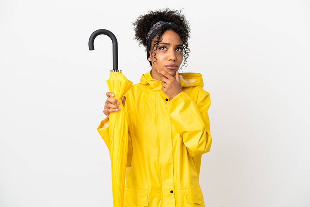 Young woman with rainproof coat and umbrella isolated on white background having doubts - Fotoğraf, Görsel