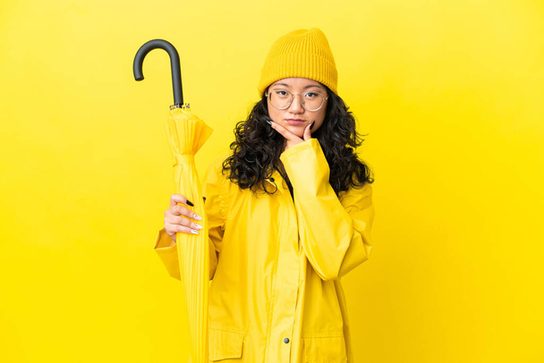 Donna asiatica con impermeabile e ombrello isolato su sfondo giallo pensando - Foto, immagini