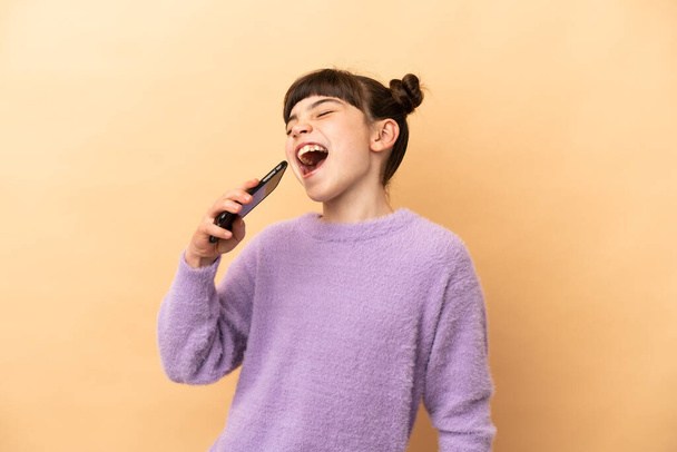 Little caucasian girl isolated on beige background using mobile phone and singing - Fotó, kép