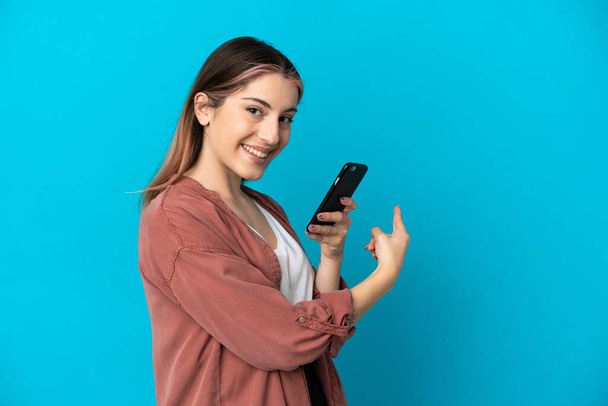 Jeune femme caucasienne isolée sur fond bleu en utilisant un téléphone mobile et pointant vers l'arrière - Photo, image