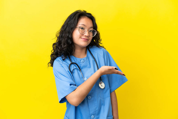 Молодий лікар хірург азіатка ізольована на жовтому тлі, представляючи ідею, дивлячись посміхаючись до
 - Фото, зображення