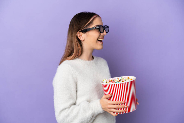 Jonge blanke vrouw geïsoleerd op blauwe achtergrond met 3d bril en met een grote emmer popcorn - Foto, afbeelding