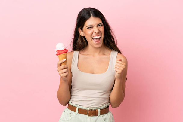 Young caucasian woman with a cornet ice cream isolated on pink background celebrating a victory in winner position - Valokuva, kuva