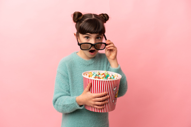 Petite fille caucasienne isolée sur fond rose surprise avec des lunettes 3D et tenant un grand seau de popcorns - Photo, image