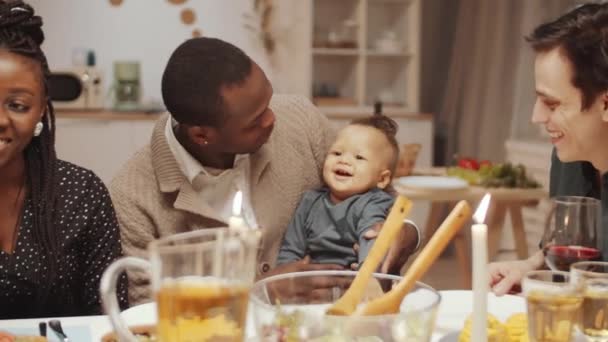 Joyeux père afro-américain assis avec un adorable bébé fils à genoux, souriant et parlant avec un enfant tout en dînant à la maison avec des amis multiethniques - Séquence, vidéo