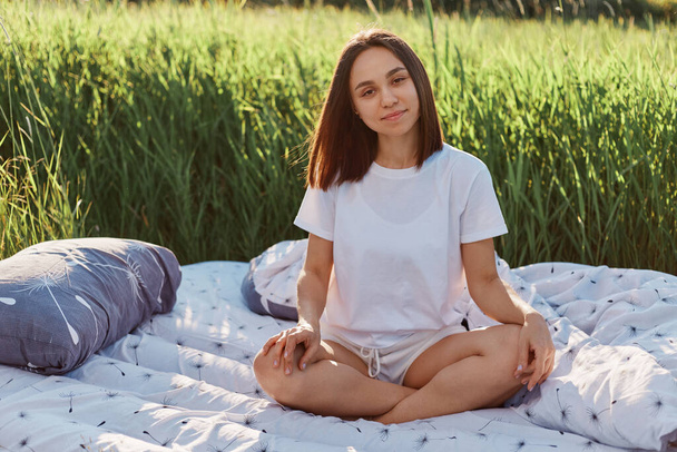 Positive attractive girl dresses white T-shirt and shorts sitting with crossed legs on bed in green field in hot sunny day on sunset, looking at camera with satisfied and calm facial expression. - Fotografie, Obrázek