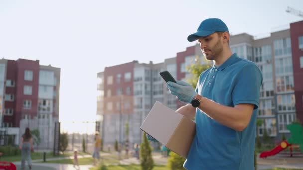 眼鏡をかけた郵便配達員が小包を運び、携帯電話で配達先を見ている。お客様の住所を検索してください。キャップと箱を持った配達人 - 映像、動画