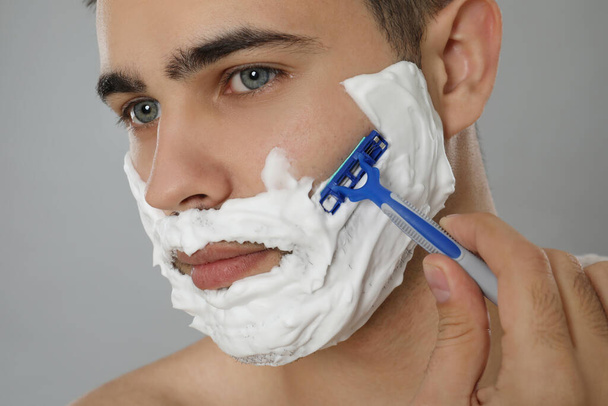 Handsome young man shaving with razor on grey background, closeup - Fotografie, Obrázek
