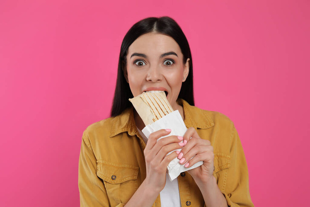 Emotional young woman eating delicious shawarma on pink background - Photo, Image