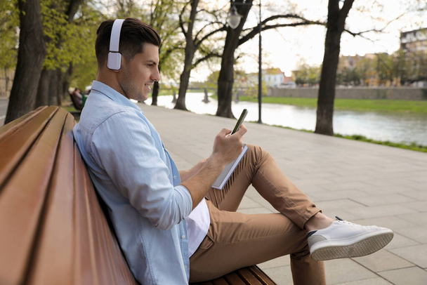 Happy man with smartphone listening to audiobook on bench near river - Photo, Image