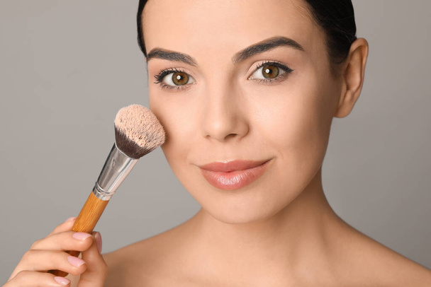Beautiful young woman applying face powder with brush on grey background, closeup - Фото, изображение