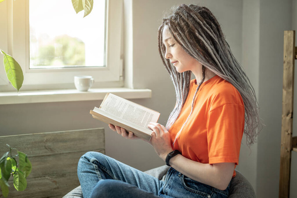 Young woman with dreadlocks and bright clothes is sitting comfortably in a bag chair and reading a book. Concept of leisure, pastime, learning and lifestyle. - Foto, imagen