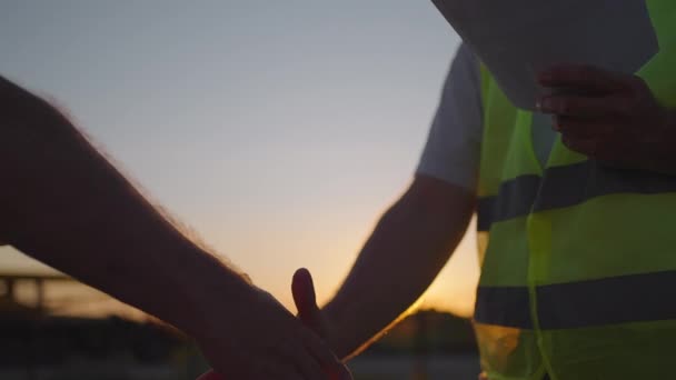 Porträt der Hände zweier Bauarbeiter. Bauunternehmer schüttelt dem Bauunternehmer auf gebautem Haus die Hand. Großaufnahme eines Handschlags zweier Männer in grünen Signalwesten vor dem Hintergrund der Sonne - Filmmaterial, Video