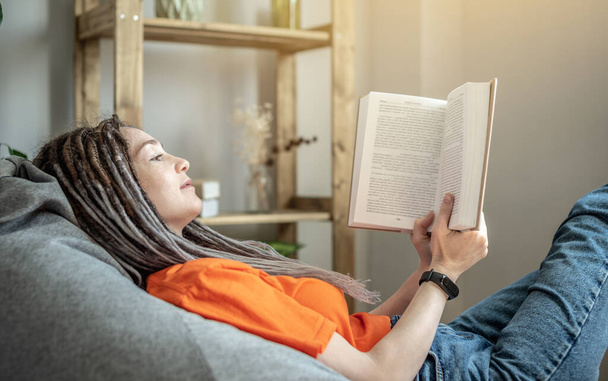 Mladá žena s dredy a světlým oblečením sedí pohodlně v křesle a čte si knihu. Koncepce volného času, zábavy, učení a životního stylu. - Fotografie, Obrázek