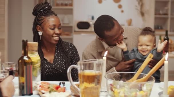 Radostný Afroameričan sedí s roztomilým synem na kolenou a usmívá se, zatímco mají sváteční večeři doma se společností multietnických přátel - Záběry, video