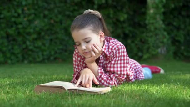 Szczęśliwa dziewczynka, w czerwonej koszuli, leżąca na zielonym dywanie, trawniku. Uśmiechnięte dziecko czyta książkę leżącą na trawie. koncepcja beztroskiego dzieciństwa, długo oczekiwane lato, letnie wakacje. - Materiał filmowy, wideo