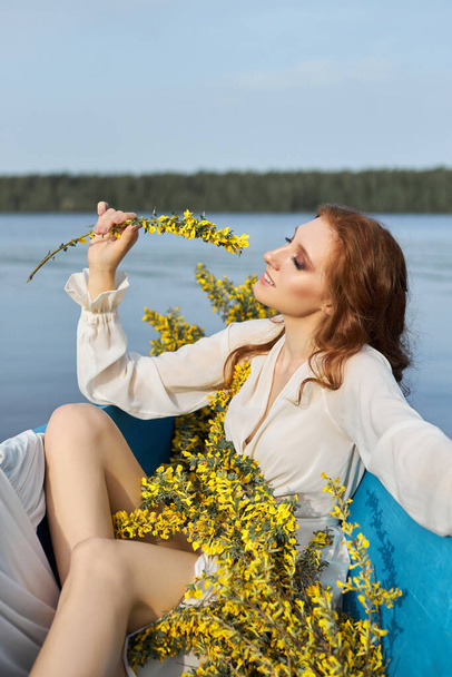 Beautiful red-haired woman lies in boat with flowers in her hands. Beauty romantic portrait girl in long white dress in wooden boat on lake - 写真・画像