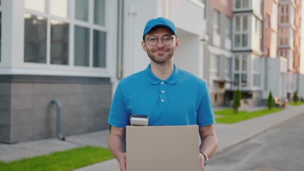 Een gelukkige bezorger met een doos in zijn handen kijkt naar de camera en glimlacht. Portret van een bezorger met een doos in slow motion - Video