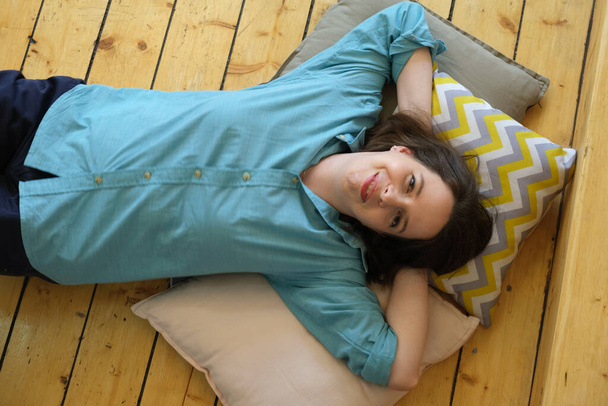 Young brunette woman with shoulder-length hair lies on pillows her head resting on her hands. Woman lies on wooden floor and smiles. Moment of rest and relaxation of a girl in a good mood. Top view - 写真・画像