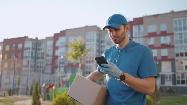 Kaukasische jonge knappe man, bezorger met blauwe pet op de straat en het dragen van kartonnen doos tijdens het gebruik van de smartphone, op zoek naar route. Mannelijke koerier met pakketafluisteren en sms 'en per telefoon. - Video