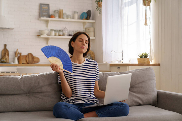 Mulher jovem sofre de insolação plana sem ar condicionado acenando ventilador sentado no sofá em casa - Foto, Imagem