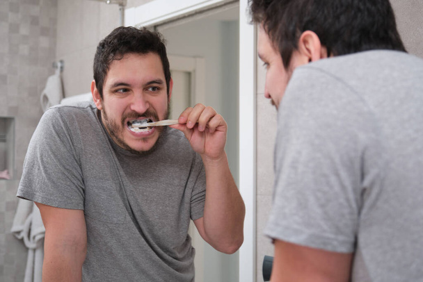Joven latino cepillándose los dientes delante del espejo en el baño por la mañana. Cepillo de dientes de bambú ecológico. - Foto, imagen